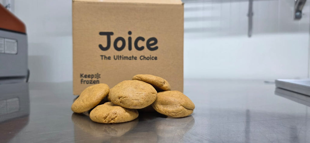 Several round frozen cookies in front of a brown cardboard box printed with the brand name 'Joice' and the phrase 'The Ultimate Choice' on a stainless steel counter.