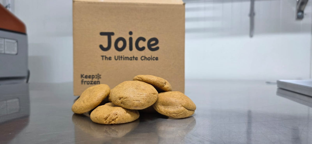 Several round frozen cookies in front of a brown cardboard box printed with the brand name 'Joice' and the phrase 'The Ultimate Choice' on a stainless steel counter.